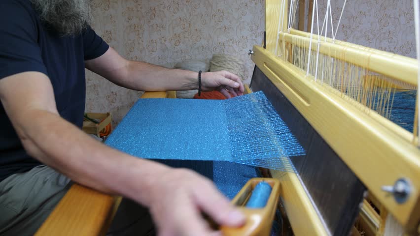 Weaver at the traditional loom. Mature man with a graying beard is weaving cloth and tapestry on a loom, using traditional techniques handicraft in the production of textile garments