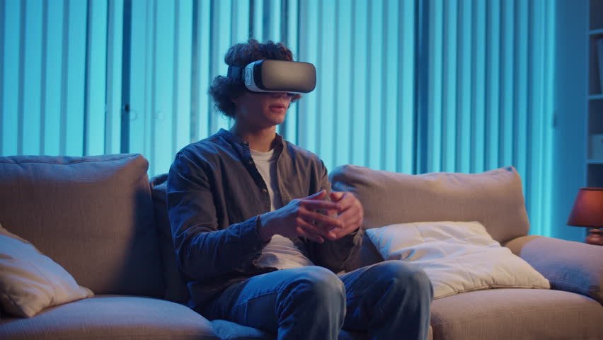 Young Man Sits on Sofa at Night in AR Glasses, Interacting with Virtual Interface. Immersive Experience in Digital World of Tomorrow. Mixed Reality, Future Technology, Mental Escape.