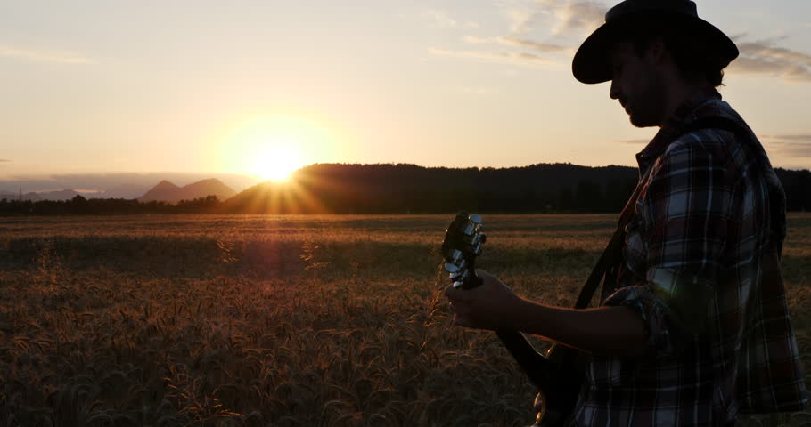 Silhouette of a musician playing guitar in a wheat field at sunset. The golden hour light casts dramatic shadows, creating a peaceful, rustic atmosphere with distant hills in the background.