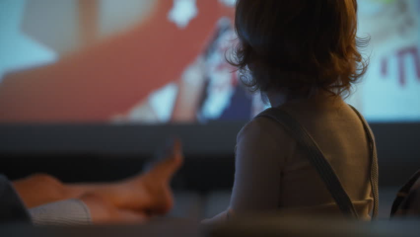 A young child is sitting on a couch, captivated by a music performance displayed on a large screen. The dim lighting adds to the cozy atmosphere, creating a perfect setting for entertainment at home