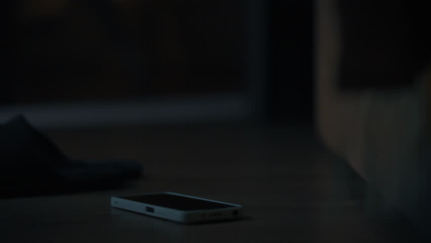In a dimly lit room, a smartphone lies on the floor beside a pair of slippers. The setting suggests a relaxed atmosphere, possibly after a long day. It is nighttime, and shadows fill the space