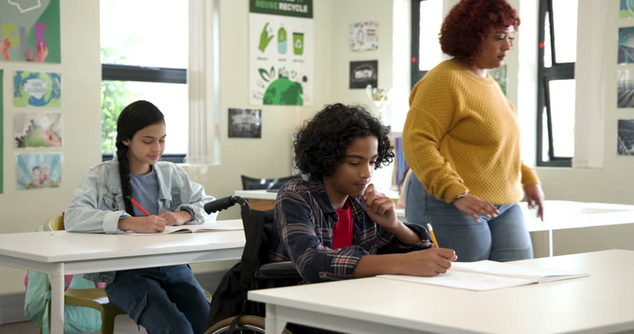 Female teacher helping boy in wheelchair with writing in classroom, fostering learning, at school. Education, diversity, inclusion, teaching, assistance, support