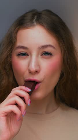 Portrait of Young Woman Eating Sweet Food as Looking at Camera. Real Person on Diet Consumes Chocolate Candy Bar as Lunch Snack. Playful Female Human Inside Cozy Living Room Alone with Sugary Fastfood