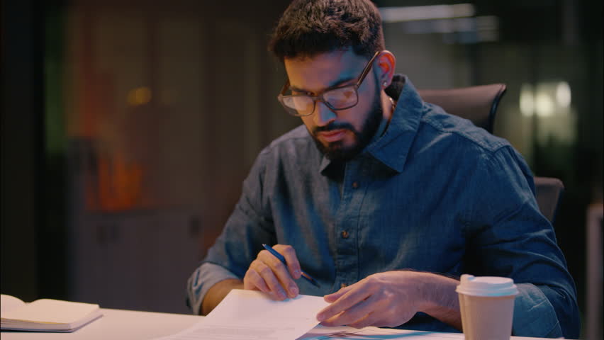 Indian ethnic man businessman reading documents paperwork on table business employer director investor lawyer sign contract deal bank insurance writing papers in evening night office overtime working