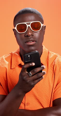 Black man, phone and sunglasses in studio, search or online shopping for fashion on orange background. Person, smartphone and trendy style with clothes, mobile app and thinking with scroll for deal