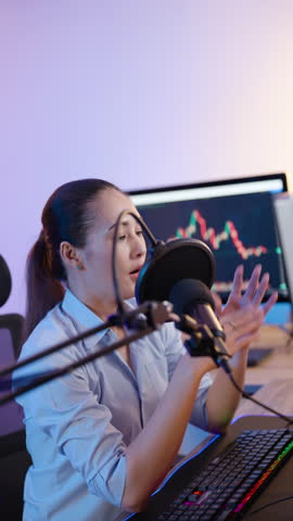 Asian woman broadcasting financial podcast with trading chart on computer screen in modern setup, focusing on stock market trends, online investment, and economic commentary in home studio.