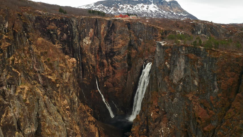Voringsfossen waterfall with powerful stream cascading down deep gorge framed by imposing barren cliffs. Distant mountain under sky in Norway