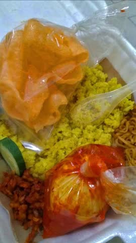 fragrant yellow nasi uduk served with noodles, sambal egg, tempeh, crackers, cucumber, and chili sauce in a takeout box, rich in flavor, color, and Indonesian street food delight.