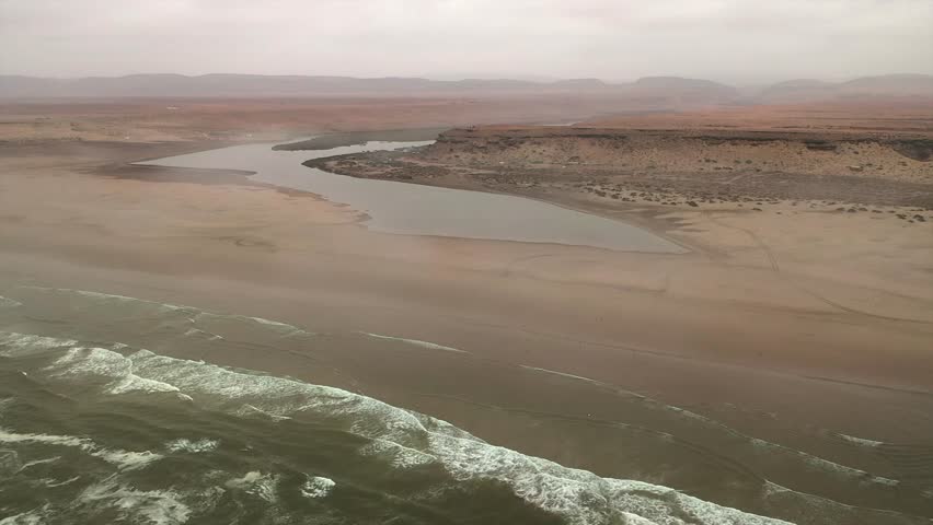 Aerial view of dramatic landscape where the desert meets the Atlantic Ocean, showing waves, sandy beach, and river mouth near Tan-Tan, Morocco