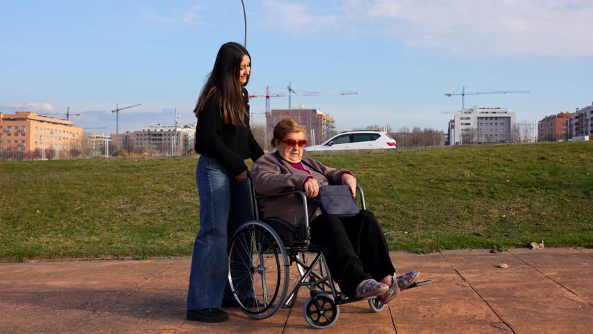 Elderly woman laughing in wheelchair outside with young woman while applying perfume on a sunny day