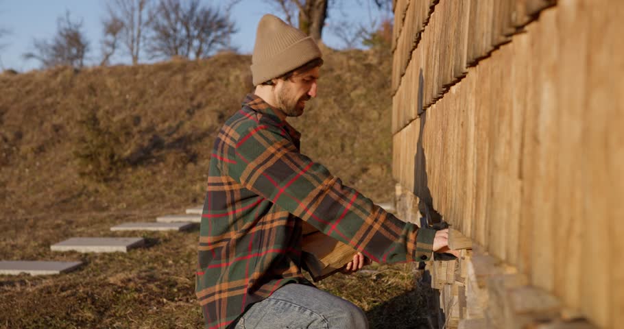 Close up of a Guy in a hat and a plaid shirt collecting firewood and walking to his country house in the evening with the rays of the sun