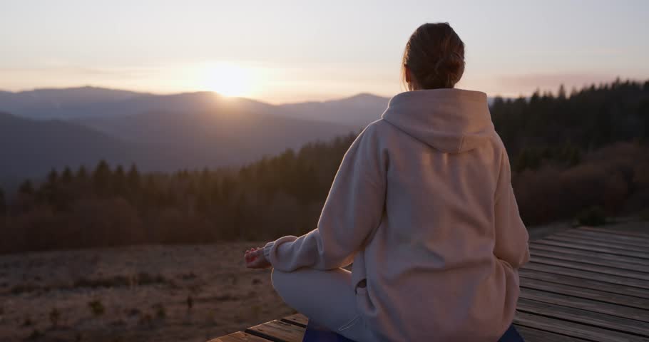 Back view of calm girl sitting on blue mat doing meditation in lotus position while watching sunset in her country house on porch among forest and mountains
