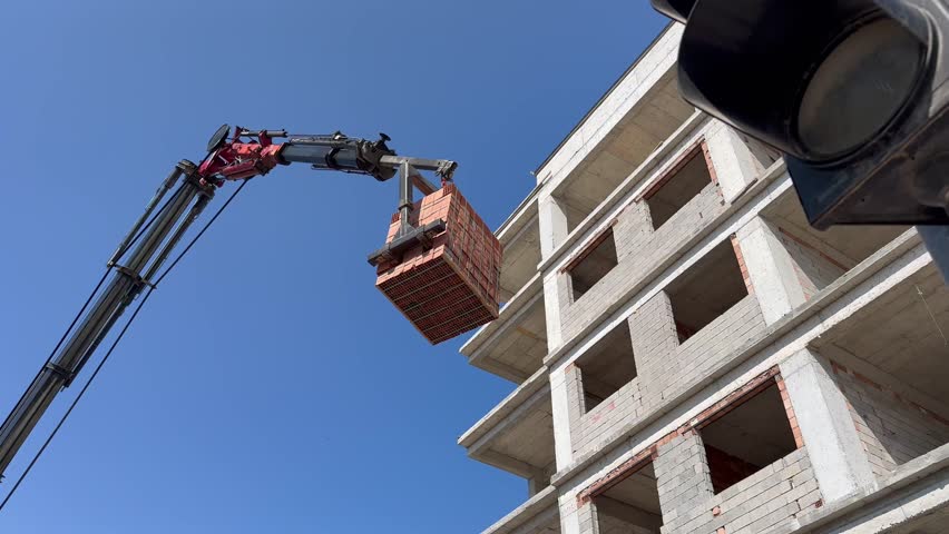 Construction site with crane lifting a pallet of bricks near an unfinished multi-story building. Concept of real estate development, urban expansion, and construction industry