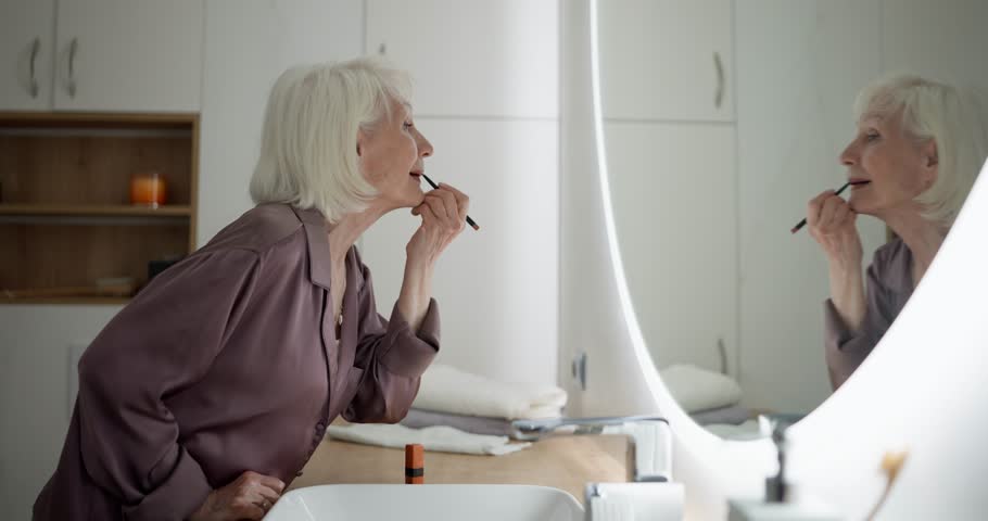 Elderly woman with white hair in purple pajamas examining her face and doing makeup in the bathroom in front of the mirror in the morning