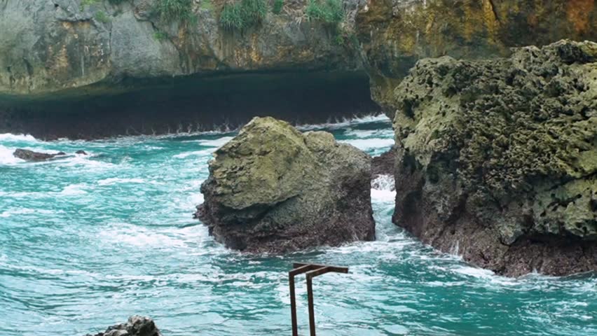 Waves crashing against rocks in tropical coastal cave in Yogyakarta, Indonesia. Powerful ocean waves hit rocky shore and cave, creating dramatic splashes and turquoise water scenery in tropical coast.
