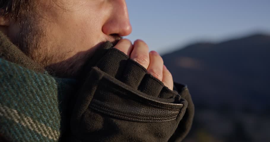 Close up of stubbled Guy warming his hands with his breath while hiking in the sunny mountains at sunset. Travel and recreation in the mountains
