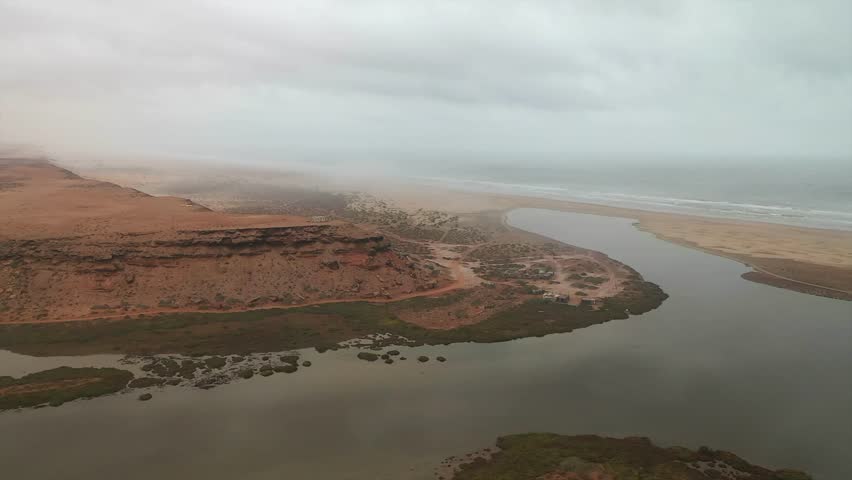 Aerial view of a desert river estuary meeting the foggy Atlantic Ocean, with a vast sandy landscape and cliffs near Tan-Tan, Morocco