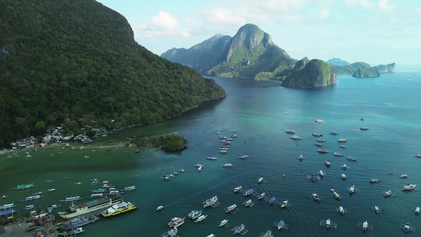 Aerial panoramic view of El Nido bay in Palawan, Philippines, with vibrant blue water, lush tropical hills, limestone islands, and numerous boats floating in the calm sea. 