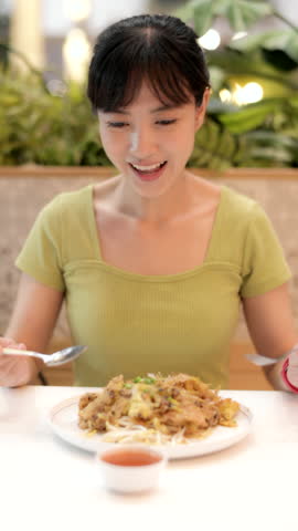 Vertical video of beautiful young asian woman with happy expression enjoying eating delicious local Thai food, crispy fried mussel pancake from bustling Bangkok shopping mall food court.