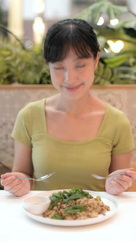 Vertical video of content Asian woman eating delicious Thai food lunch in Bangkok restaurant, enjoying local basil stir fry with rice. tasty and authentic food lifestyle moment.