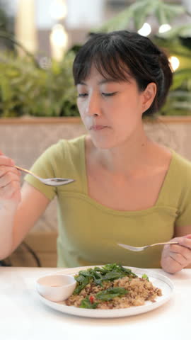 Vertical video of content Asian woman eating delicious Thai food lunch in Bangkok restaurant, enjoying local basil stir fry with rice. tasty and authentic food lifestyle moment.