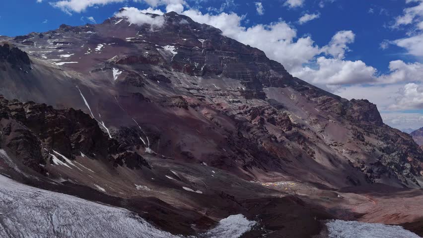 Aerial view of Aconcagua, Argentina, with its rugged slopes and glaciers. The base camp with colorful tents is visible below, contrasting against the harsh Andean terrain.