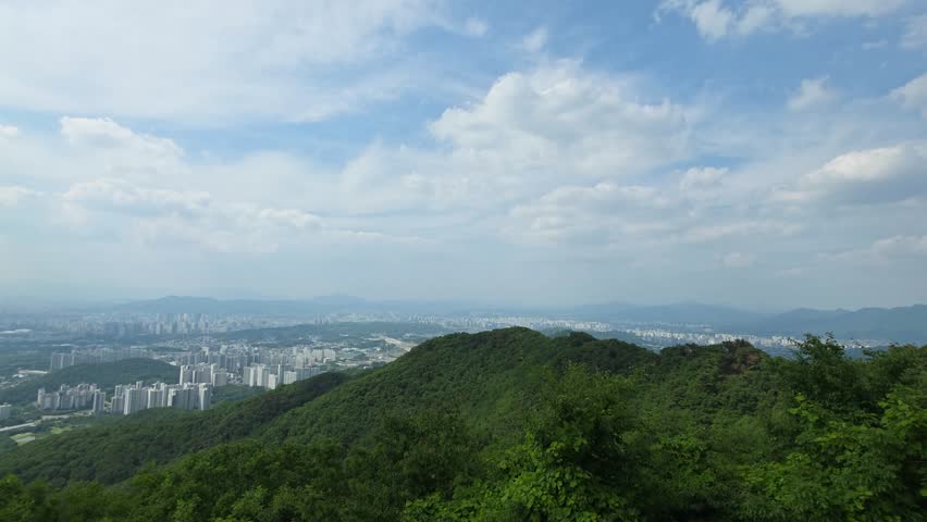 A stunning timelapse from Namhansanseong Mountain shows clouds drifting across a blue sky above the sprawling Seoul cityscape, with lush green forest covering the mountainside in the foreground.