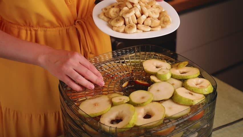 Housewife adding dried banana slices to a food dehydrator filled with various sliced fruits