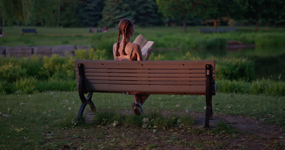 A young woman reads quietly on a park bench beside a tranquil pond. A slow dolly movement captures the serenity, focus, and emotional depth of the moment at golden hour.