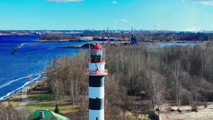 A striped lighthouse stands on the riverbank near leafless trees and industrial cranes, marking the entrance to a port where land meets water under a clear sky.