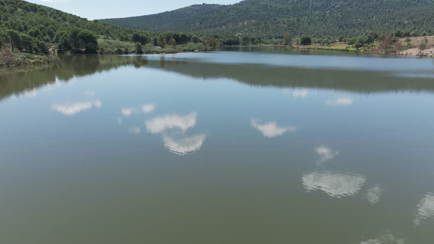 Low side drone shot of a vibrant lake in Badajoz, Spain, with blue-green water, wildflowers, diverse vegetation, and forested hills under a blue sky with scattered white clouds reflecting in the water