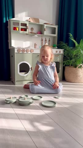 young child smiles while playing with kitchen toys in well-decorated, light-filled room. The miniature kitchen features a variety of utensils and colorful items adding charm