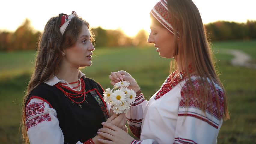 Two girls in traditional embroidered clothing hold a bunch of daisies at sunset, one gently picking petals while the other watches, evoking a scene of playful fortune telling.