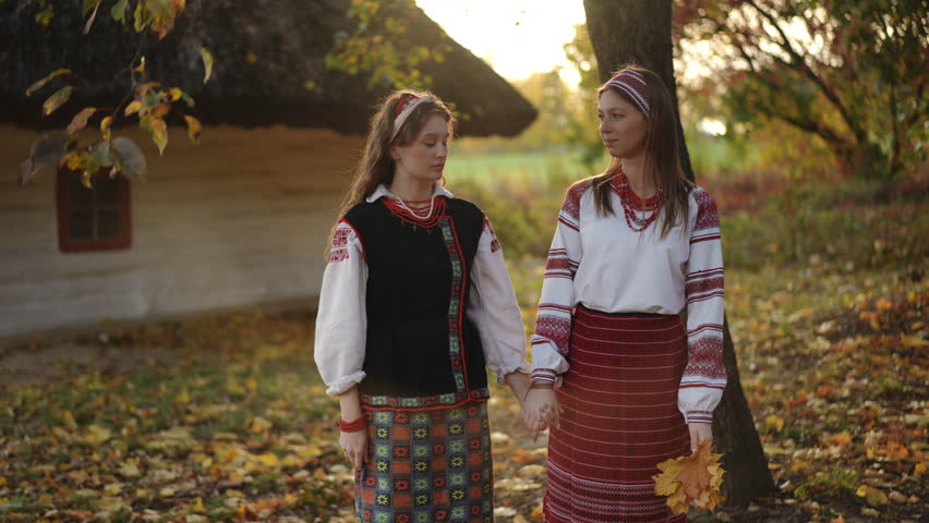 Two young women in traditional Ukrainian outfits walk hand in hand near a thatched house, surrounded by colorful autumn leaves, captured in slow motion with warm sunset light.