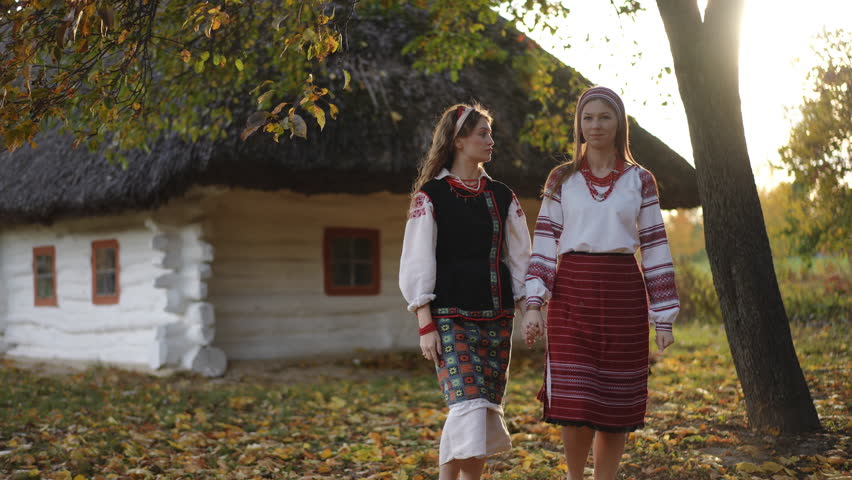 Two girls in authentic Ukrainian folk costumes walk hand in hand near a whitewashed thatched house, surrounded by autumn foliage and golden evening light, captured in slow motion.