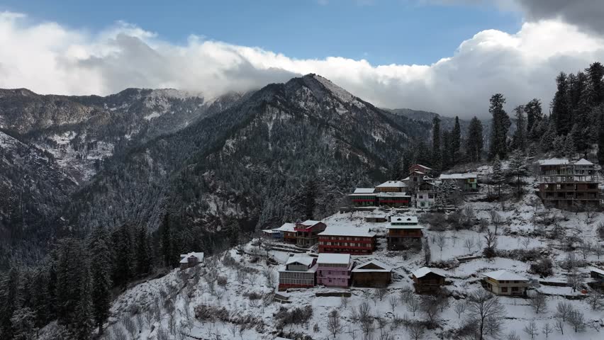 Sunlight breaks through after a snowfall, revealing a Himachali village nestled in snow-covered hills under a bright blue sky.