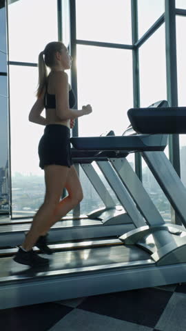 Portrait of young blonde woman runs on modern treadmill near panoramic window side view. Slim athletic lady exercises with trainer machine in luxury gym slow motion. Vertical shot