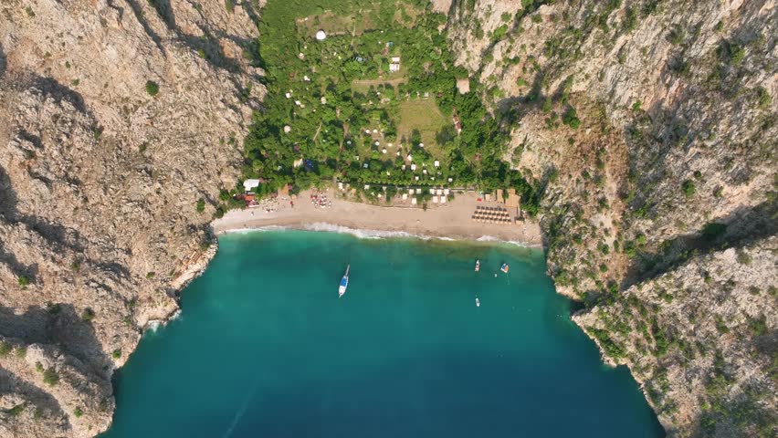 Butterfly valley, a stunning natural wonder, reveals its turquoise waters, anchored boats, beach umbrellas dotting the sandy shore, and lush green vegetation clinging to the steep cliffs