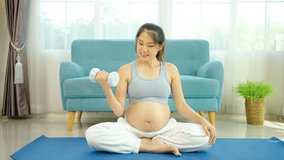 Young pregnant Asian woman about to become a mother, wearing a sports bra, exercising, and sitting lifting dumbbells up and down slowly to flex her arm muscles before giving birth on a yoga mat - Powered by Shutterstock - Get 15% off with code: PIKWIZARD15