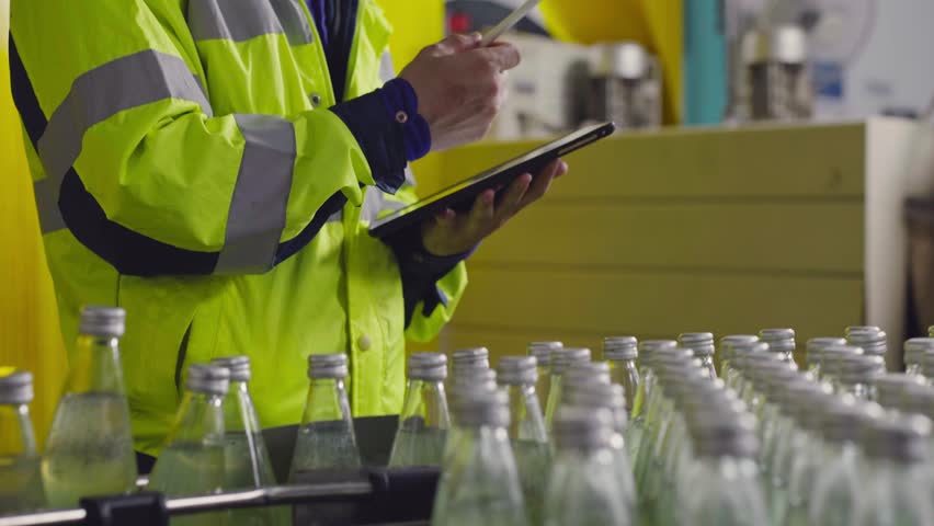 A male factory worker writing on digital tablet inspecting quality of juice bottle on conveyor belt at drink industry. Inspector working represents for responsibility and safety