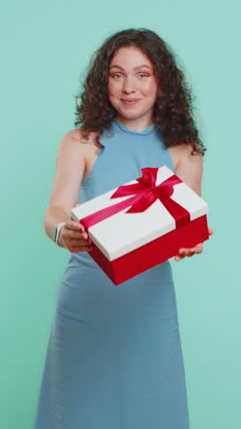 Positive smiling young woman presenting birthday green gift box stretches out hands, offer wrapped present career bonus, celebrating party sale. Girl isolated alone on blue studio background. Vertical