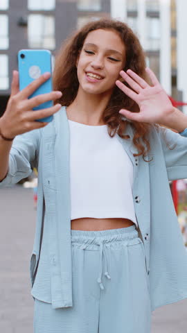 Pretty preteen child kid blogger taking selfie on smartphone, communicating video call online with subscribers recording stories for social media vlog. Teen girl walking in urban city street. Vertical