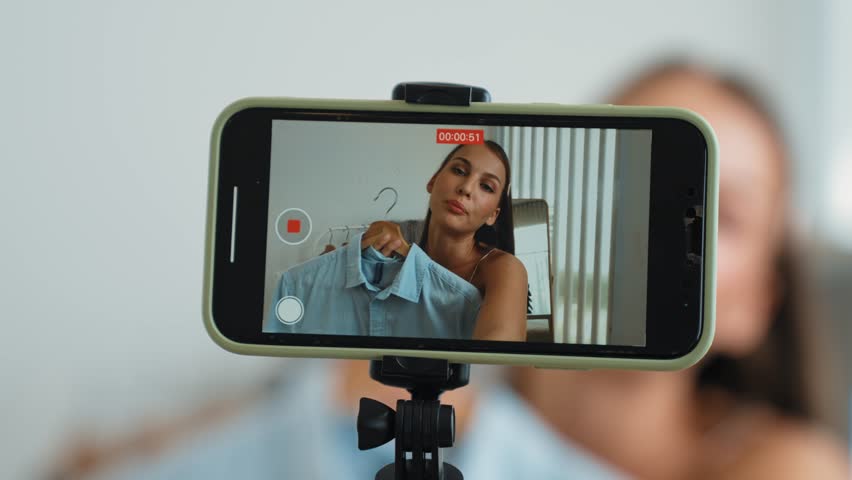Young social medial content creator woman make fashion video. Blogger smiles to camera using selfie stick and light ring while making persuasive online clothing sell vlog to audience or follower. Adit