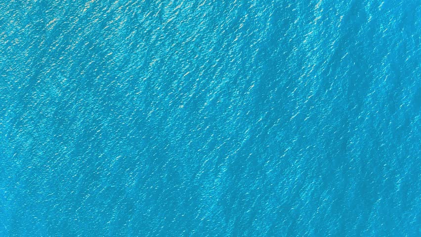 Light blue background of sea water and reflections of the sun on the waves. Drone flying over the ocean. Waves of the water surface. Flying over the sea. Sea sunrise, ripples of the ocean waves.