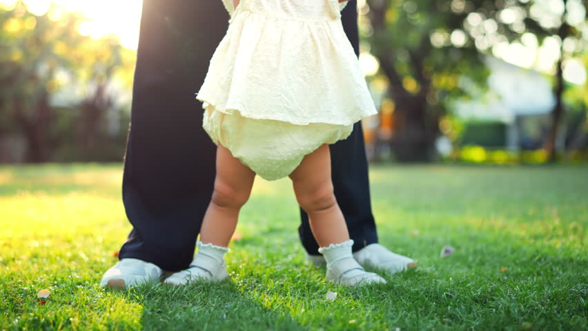 Close-up of baby wearing shoes taking first steps on green grass in park with soft morning sunlight – toddler walking with mother, childhood milestone and outdoor lifestyle concept