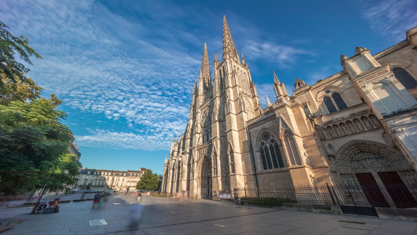Cathedrale Saint-Andre de Bordeaux timelapse hyperlapse with twin spires under a vibrant blue cloudy sky. People walking in the square create a serene urban atmosphere before sunset. Bordeaux, France