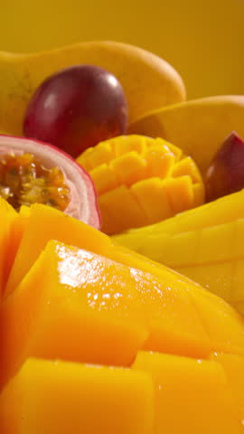 Tropical fruits including juicy sliced mango passion fruit and whole ripe fruits in a vivid yellow setting. Freshness, vibrant color and summer vibes. Vertical close-up slow motion