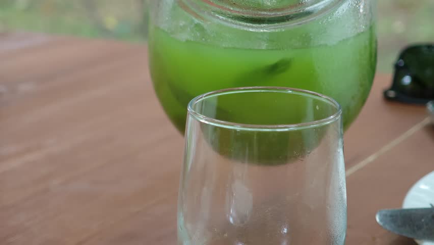 Green juice being poured into a glass with ice on a wooden table