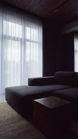 Dark modular sofa resting near tall glazed doors veiled by curtains. Gentle backlight crafts contemplative ambience suitable for cinematic B‑roll or design moodboards.