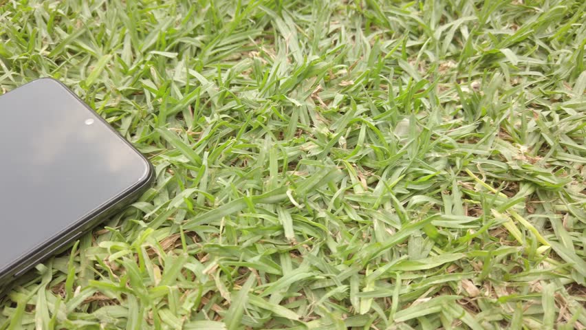 Modern black smartphone lying on green grass reflecting the sky, with slow pan showing reflection and nature contrast.